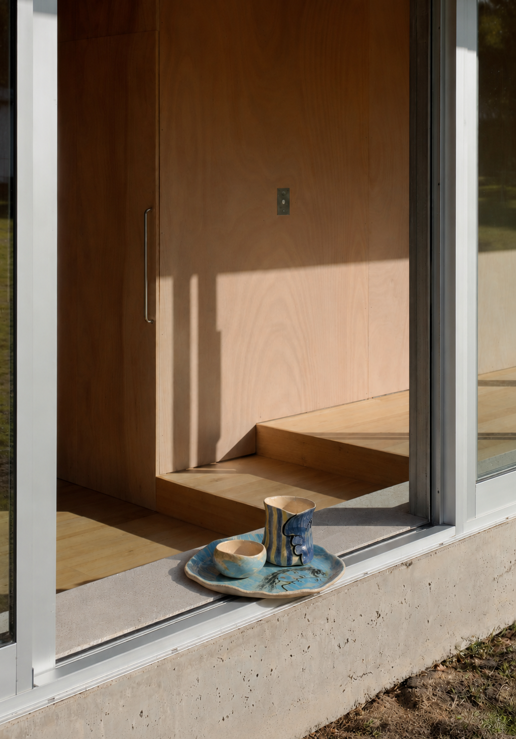 Plat au visage bleu posé sur un seuil de béton devant une baie vitrée ouverte sur un intérieur en bois